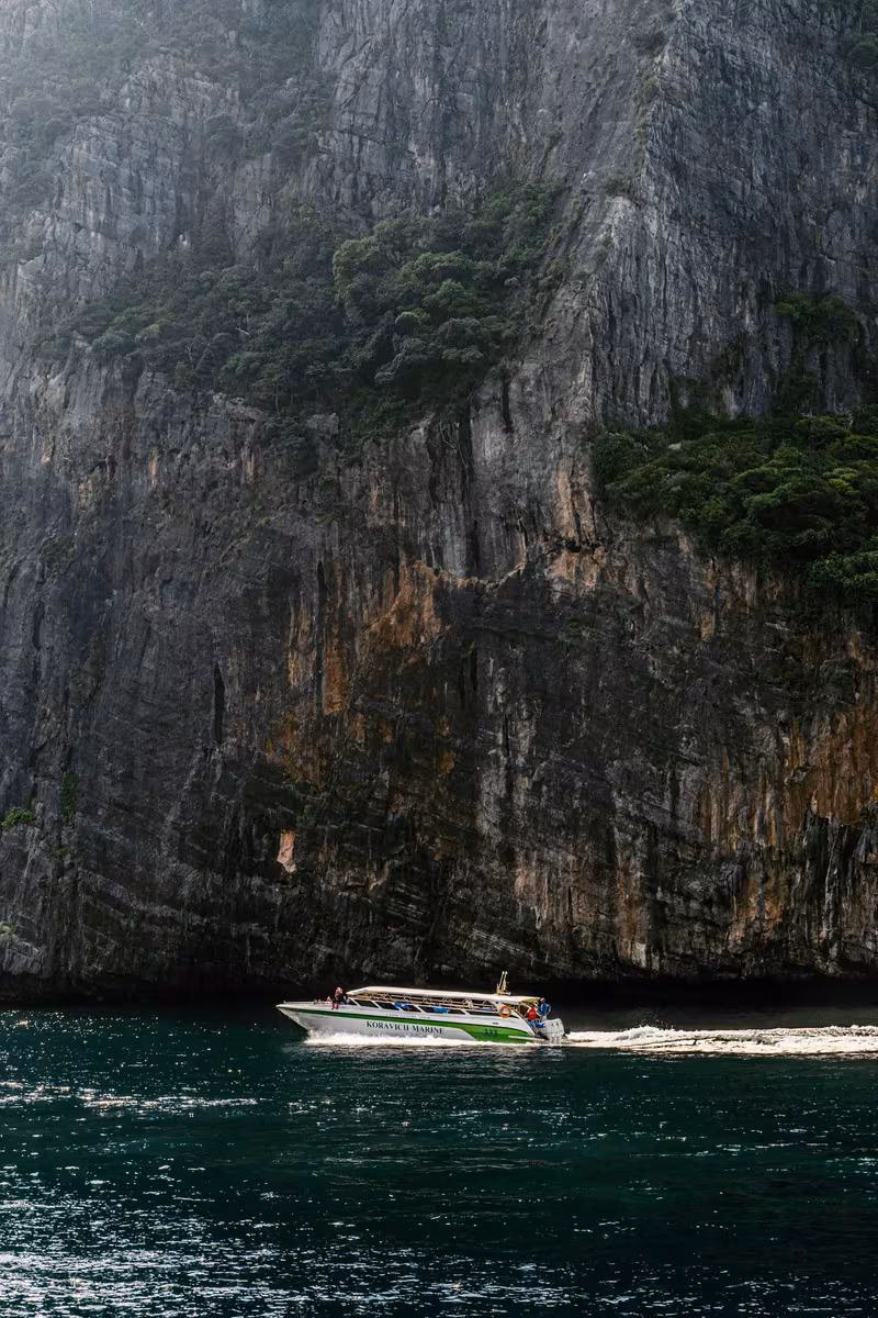 Thailand: Motorboot fährt vor einer steilen, bewaldeten Felswand auf dunklem Wasser.