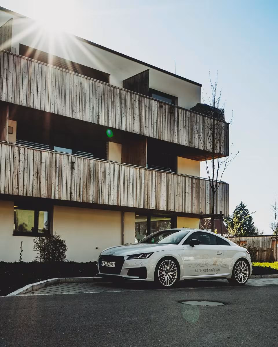 Weißer Audi vor modernem Holzhaus mit aufsteigender Sonne.