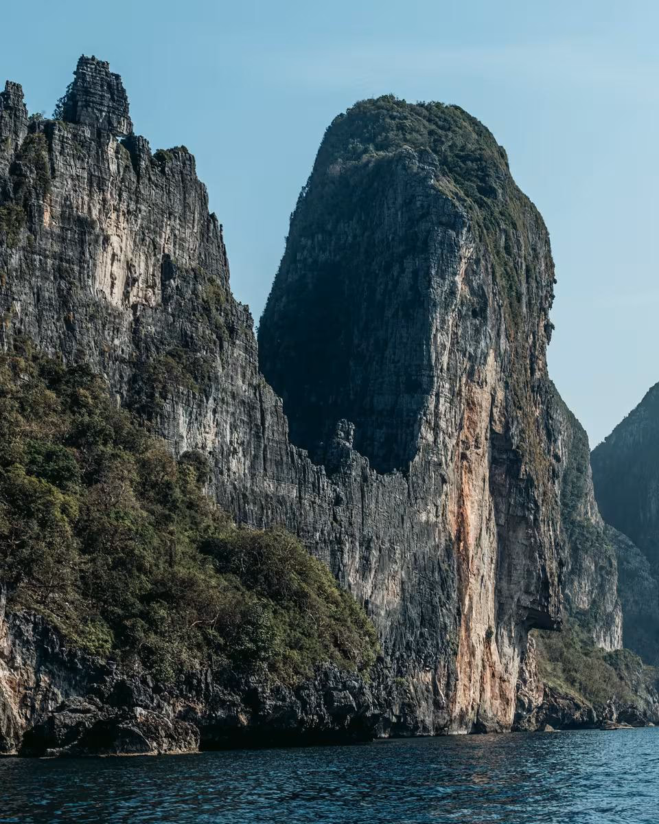 Thailand: Steile Kalksteinklippen mit Vegetation am Meer unter blauem Himmel.