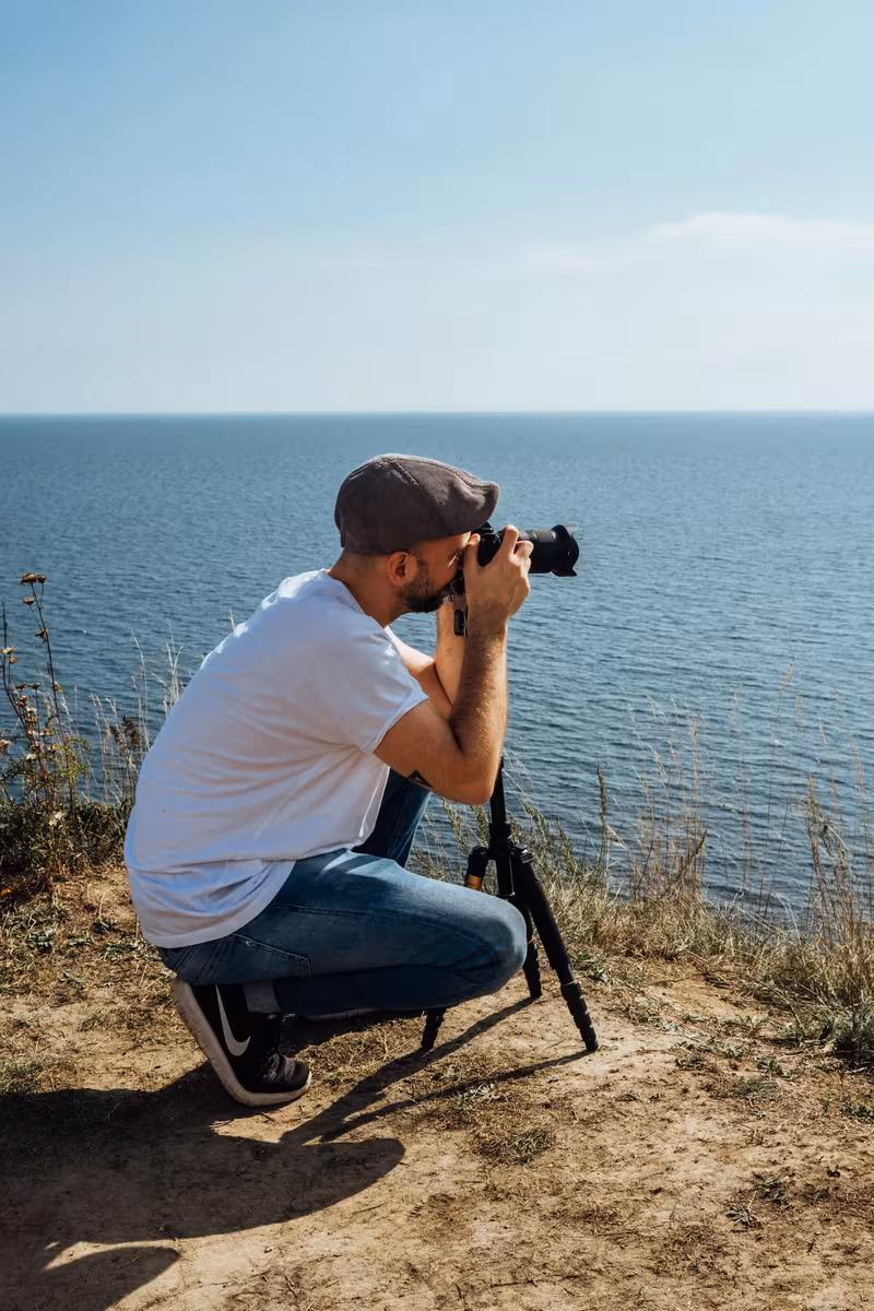 Copenhagen: Mann mit Kamera und Stativ kniet an einer Klippe vor dem Meer.