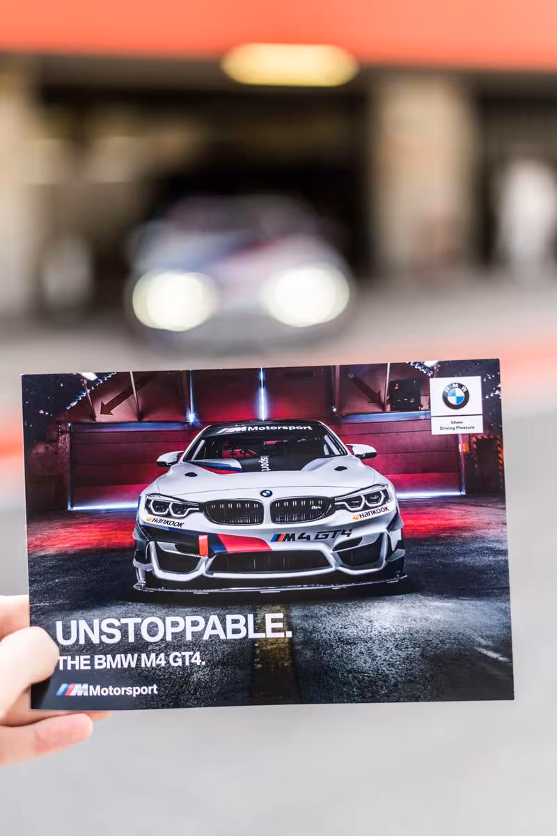 BMW Motorsport: Hand hält Plakat mit BMW M4 GT4 und Schriftzug „UNSTOPPABLE“ vor unscharfem Rennstrecken-Hintergrund.