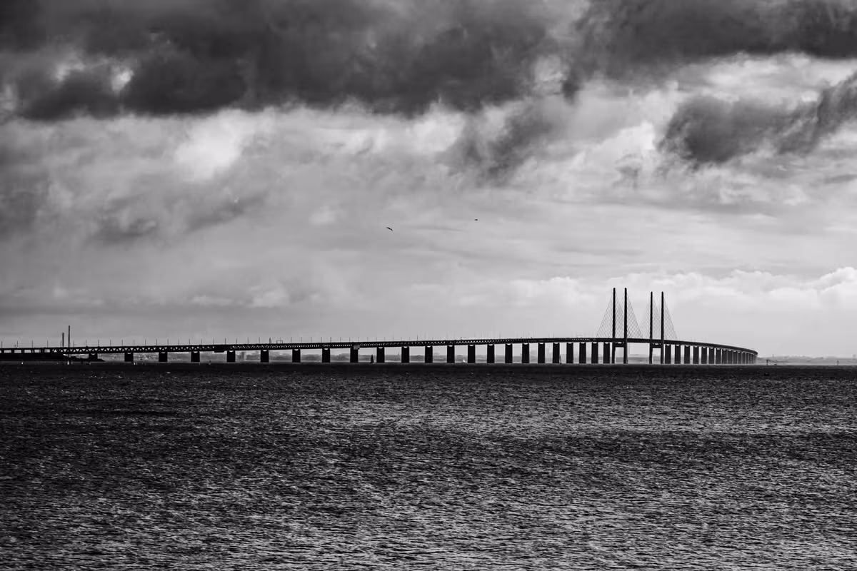 Copenhagen: Schwarz-weißes Foto einer langen Brücke mit Pylonen über dem Meer unter bewölktem Himmel.