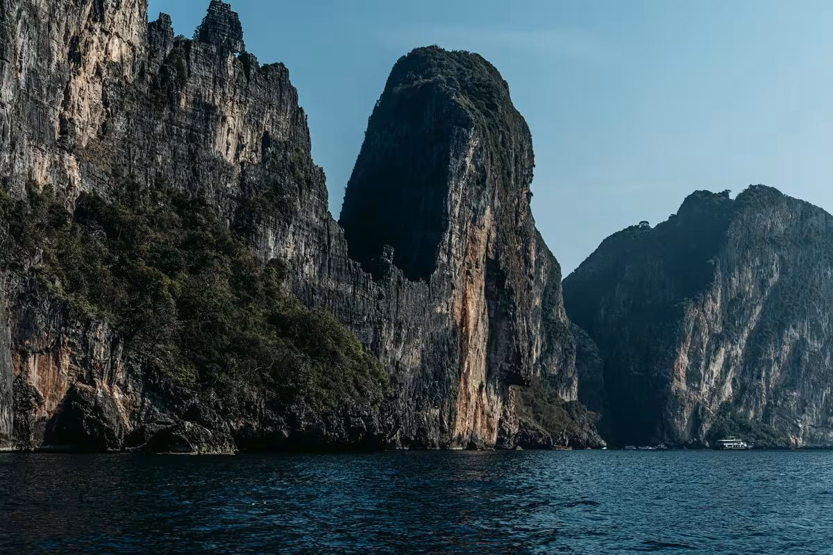 Thailand: Steile, bewachsene Felsklippen ragen aus dem ruhigen Meer unter einem klaren Himmel.