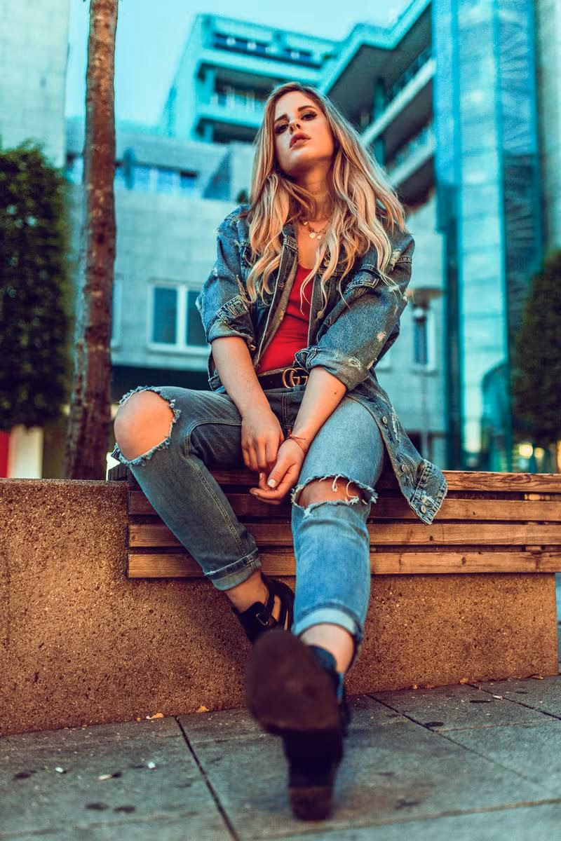 Lifestyle: Blonde Frau in Jeansjacke und rotem Top sitzt auf einer Holzbank im urbanen Raum.
