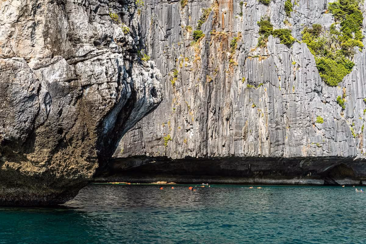 Thailand: Steile Kalkfelsenklippe mit Felsenhöhle und türkisfarbenem Meer im Vordergrund.