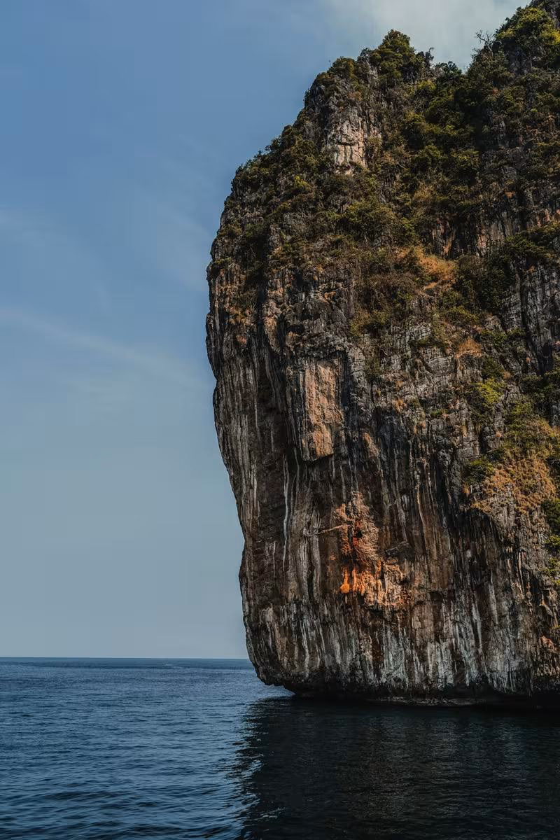 Thailand: Steile, bewachsene Klippe, die senkrecht in ein ruhiges Meer ragt.