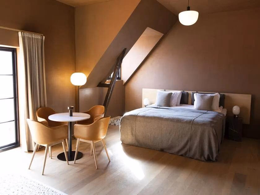 Axor: Minimalistisches Schlafzimmer mit Bett, Tisch und Stühlen unter schräger Decke.