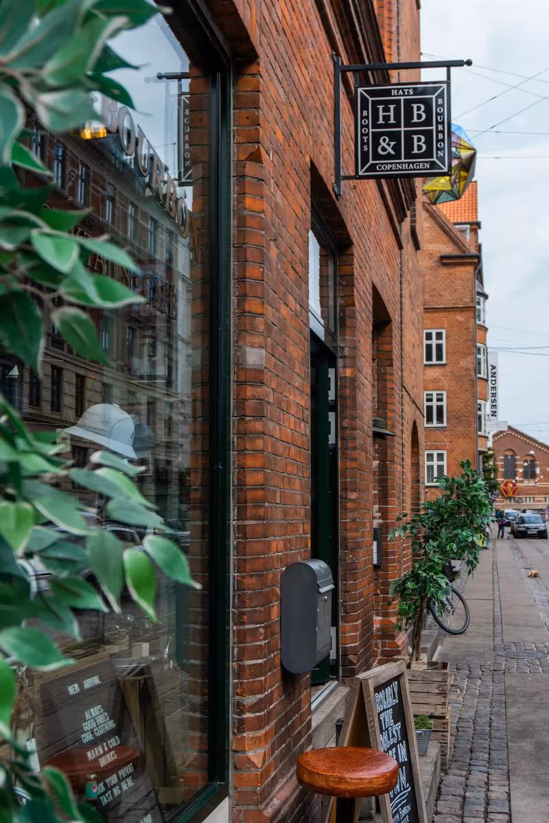 Copenhagen: Ziegelgebäude mit Schaufenster, Hängeschild „Hats, Boots & Bourbon“ und Pflanze im Vordergrund.