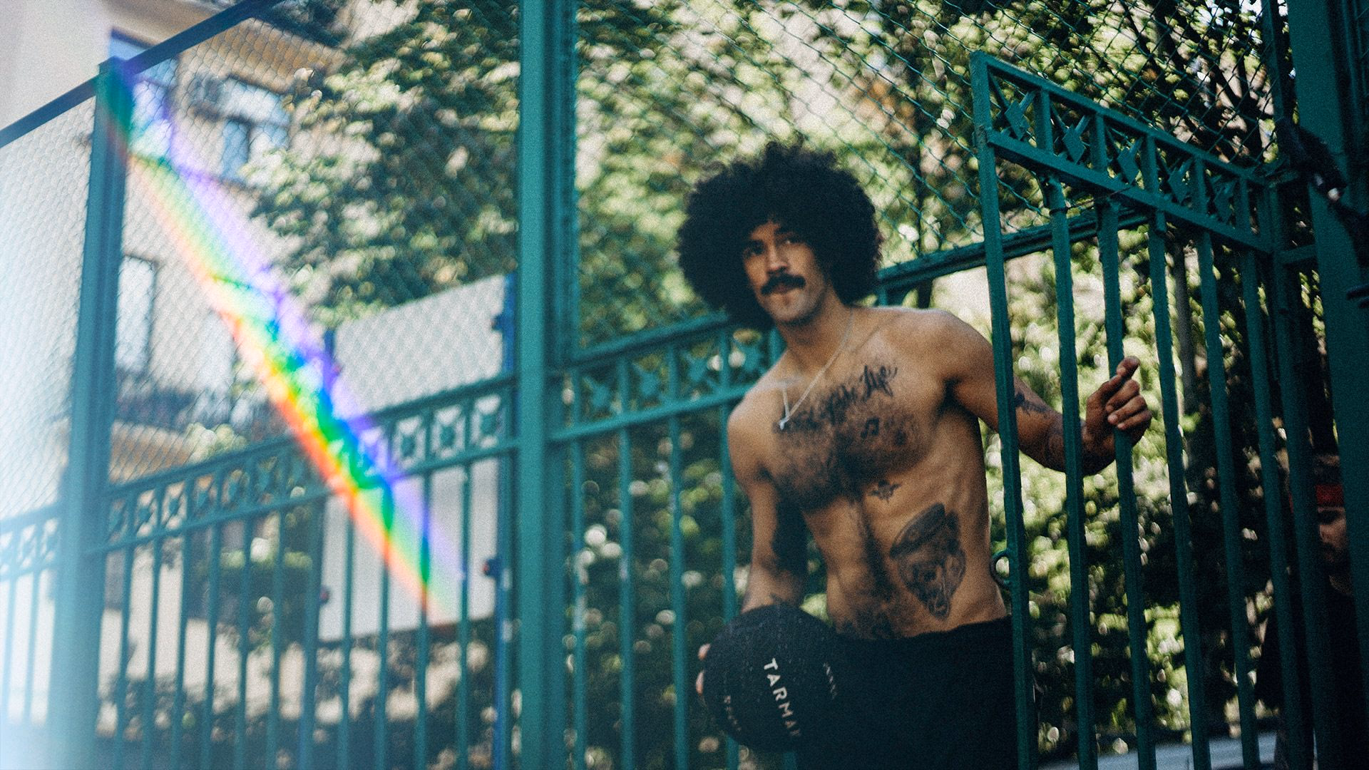 Bärtiger Mann mit Afro und Tattoos hält eine schwarze Kappe vor grünem Zaun.