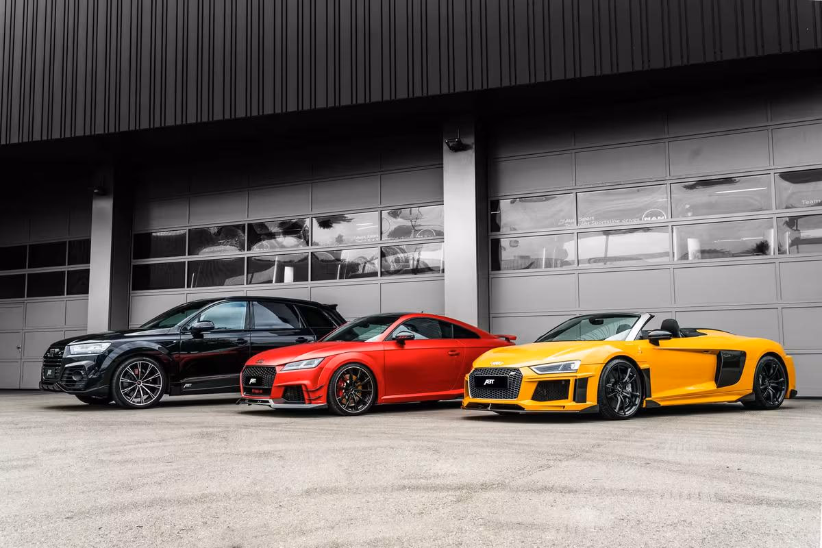 Drei Audi-Modelle in Schwarz, Rot und Gelb vor einer modernen Industriehalle.