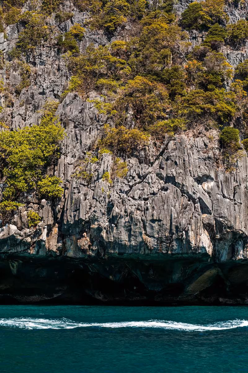 Thailand: Steile, bewachsene Felsklippe mit Höhle über türkisfarbenem Meerwasser.