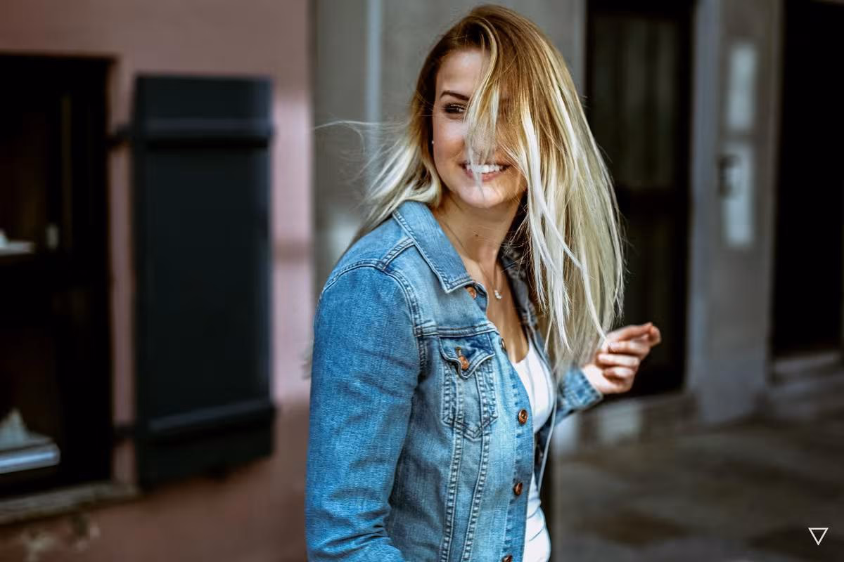 Lifestyle: Lachende blonde Frau in Jeansjacke vor einem Haus mit rosafarbener Fassade.