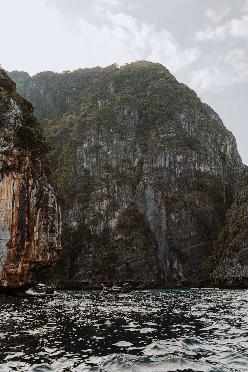 Thailand: Langschwimmboote vor steilen, bewaldeten Kalkfelsen unter bewölktem Himmel