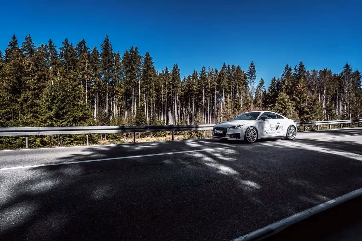 Silberner Audi auf Landstraße vor Nadelwald unter blauem Himmel.