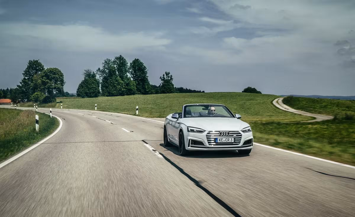 Weißer Audi Cabriolet fährt auf einer Landstraße durch eine grüne Landschaft.