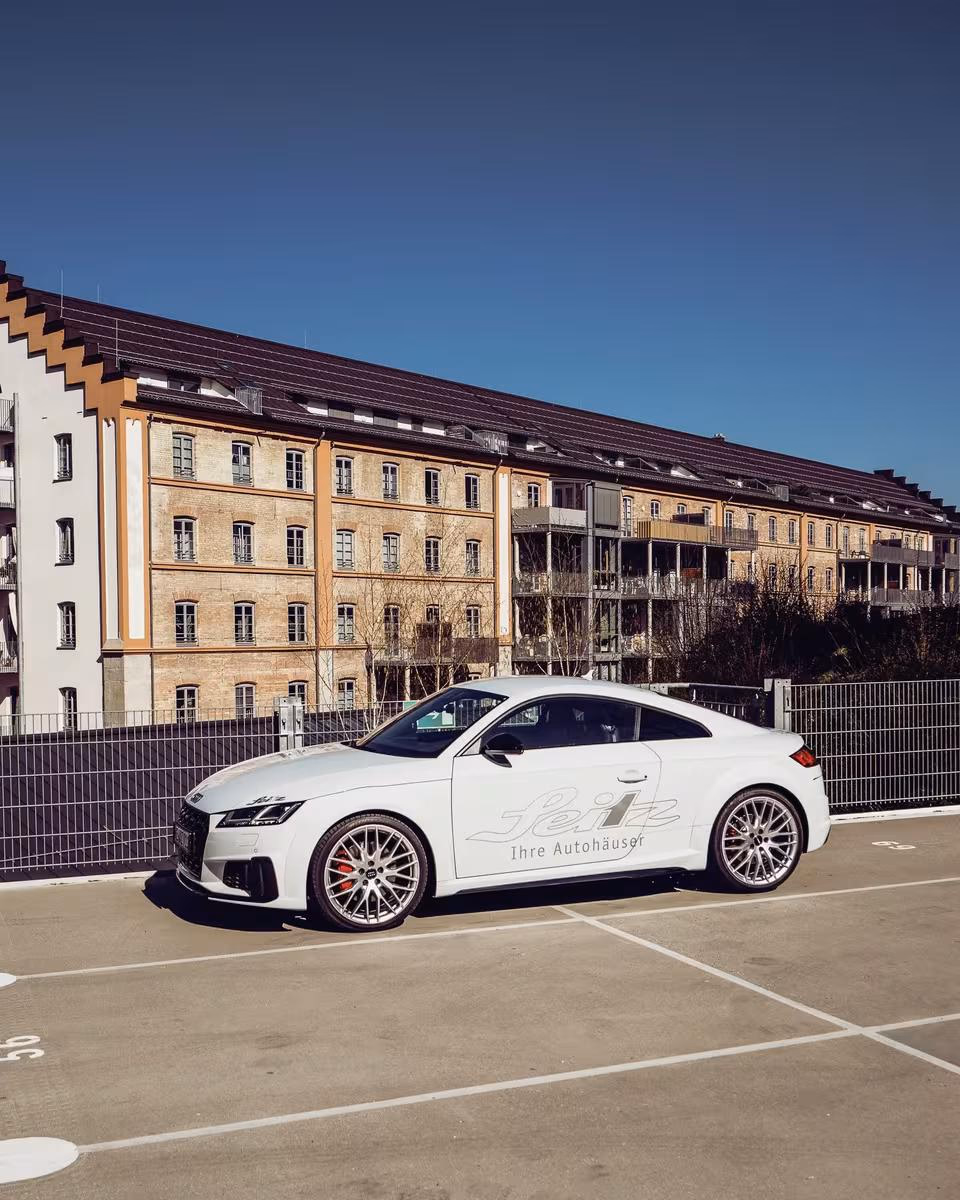 Weißer Audi RS5 Coupé mit „Seitz“ und „Ihre Autohäuser“-Schriftzug vor einem Gebäude.