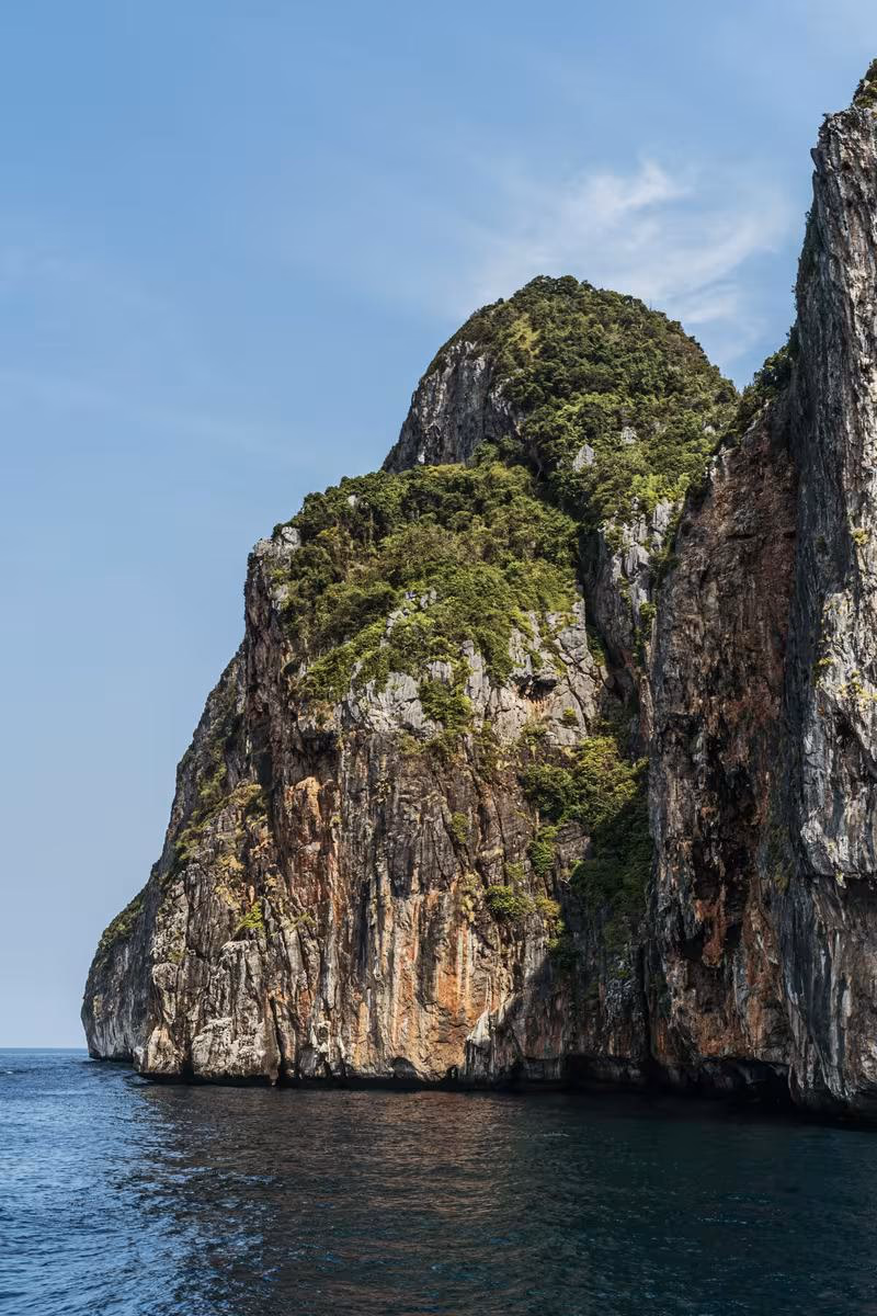 Thailand: Steile, bewachsene Felsklippe, die direkt ins Meer ragt, unter blauem Himmel.