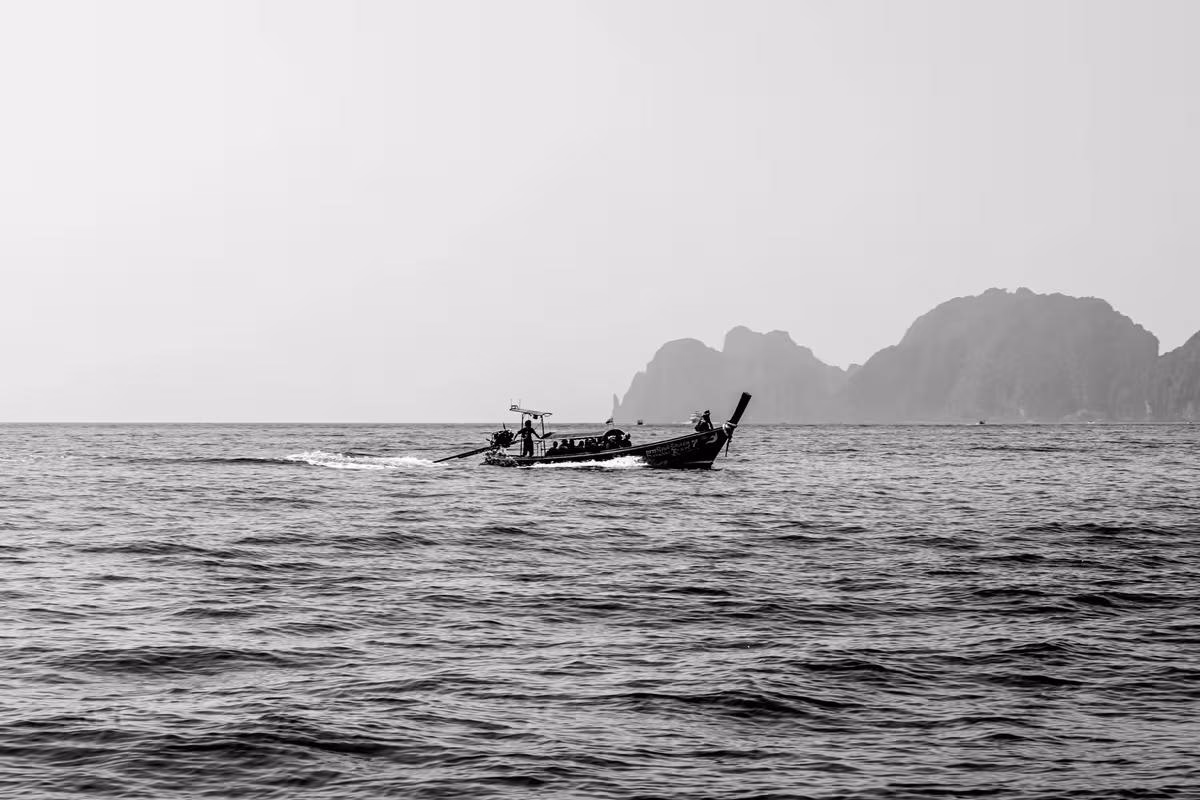 Thailand: Schwarzes Langschwimmboot mit Passagieren auf dem Meer vor felsigen Klippen.