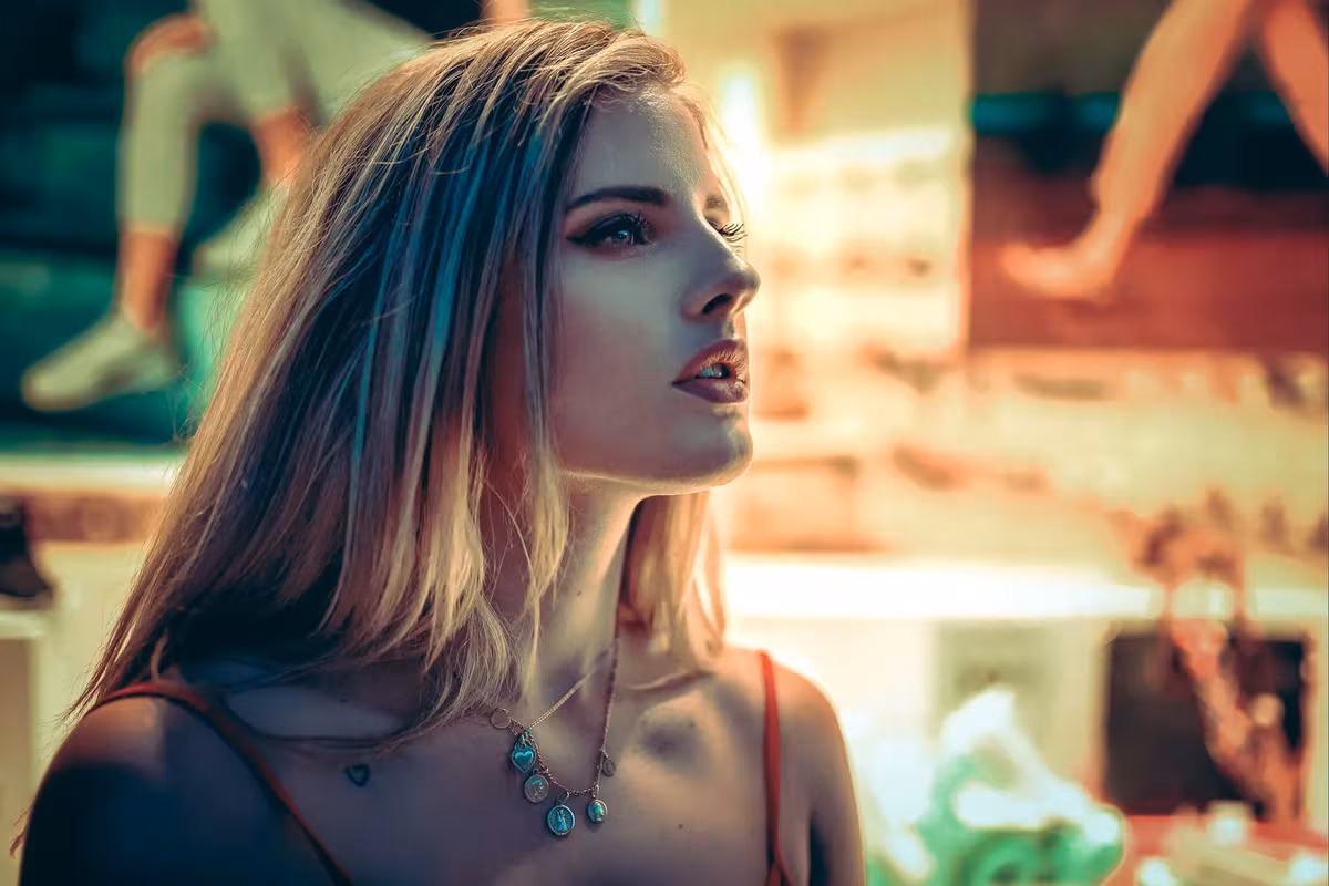 Lifestyle: Blonde Frau im Profil mit Schmuck vor unscharfem, farbigem Hintergrund.