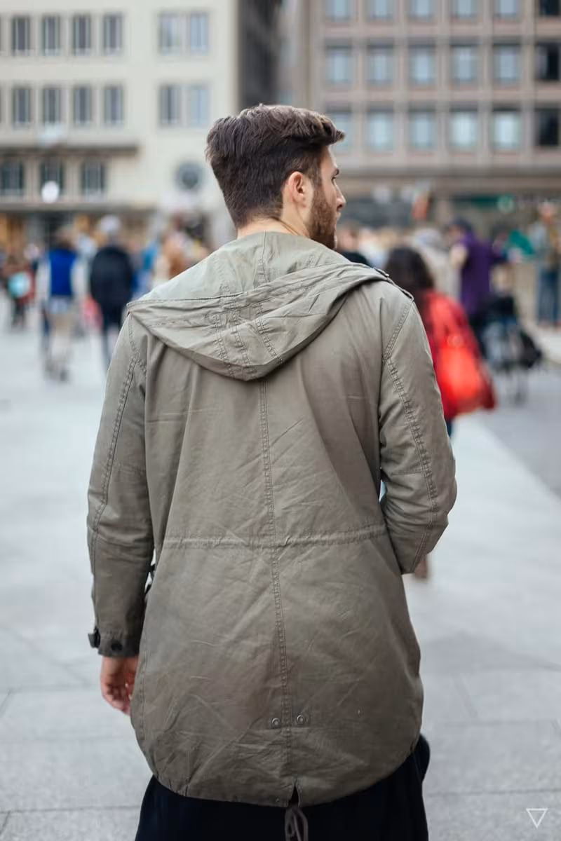 Lifestyle: Mann mit olivgrüner Kapuzenjacke von hinten auf einem belebten Stadtplatz.