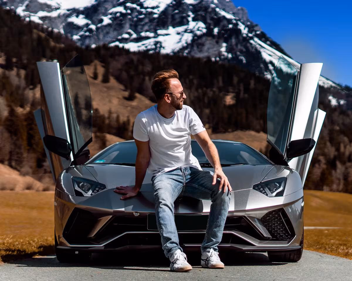 Lifestyle: Mann sitzt lässig auf der Motorhaube eines silbernen Lamborghini vor verschneiten Bergen.
