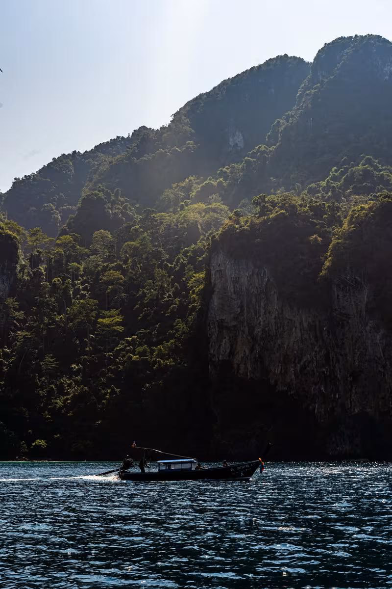 Thailand: Langschwimmboot fährt vor steilen, bewaldeten Felsklippen über ruhiges Meer.