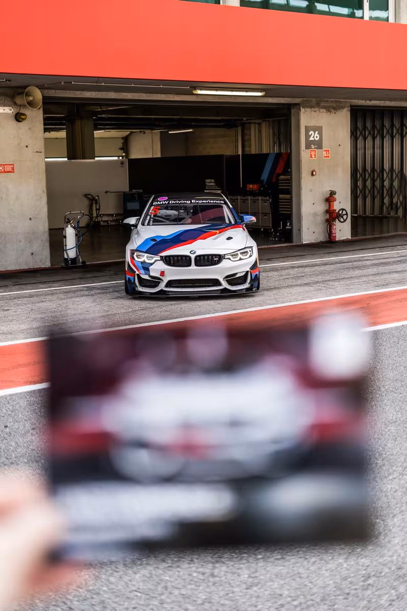 BMW M4 Motorsport-Rennwagen mit M-Farben vor Boxengasse, davor unscharfes Handy.