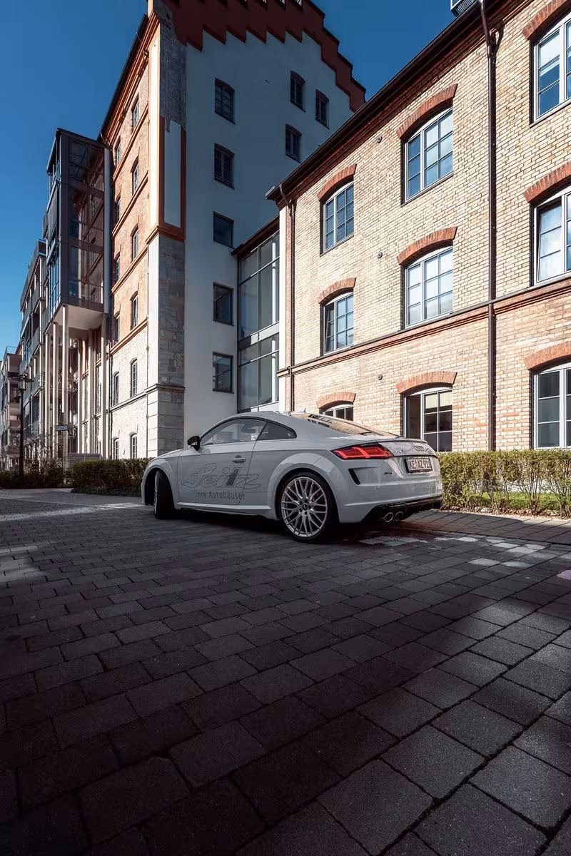 Silberner Audi TT mit „Jeiz“-Aufkleber vor einem Backstein- und Glasgebäude.