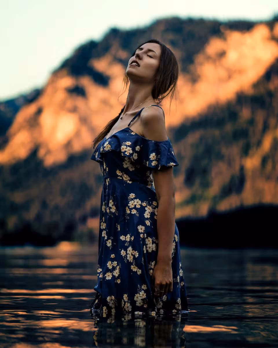 Lifestyle: Frau im blauen Blumendress steht im See vor sonnenbeschienenen Bergen.