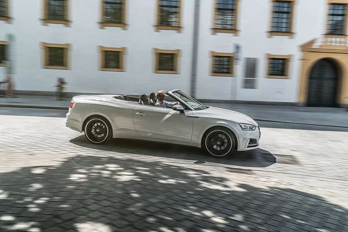 Silberner Audi Cabriolet fährt auf Kopfsteinpflaster vor weißem Gebäude.