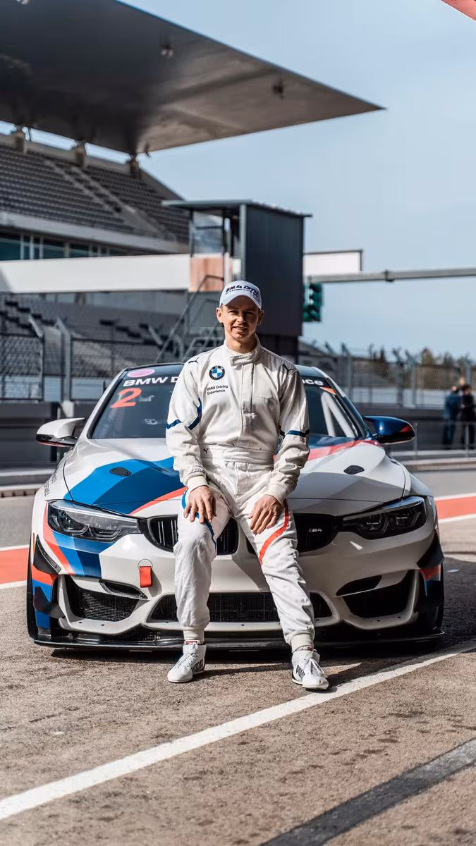 BMW Motorsport: Rennfahrer im weißen Overalls sitzt lässig auf der Motorhaube eines BMW M4 GT3 auf der Strecke.