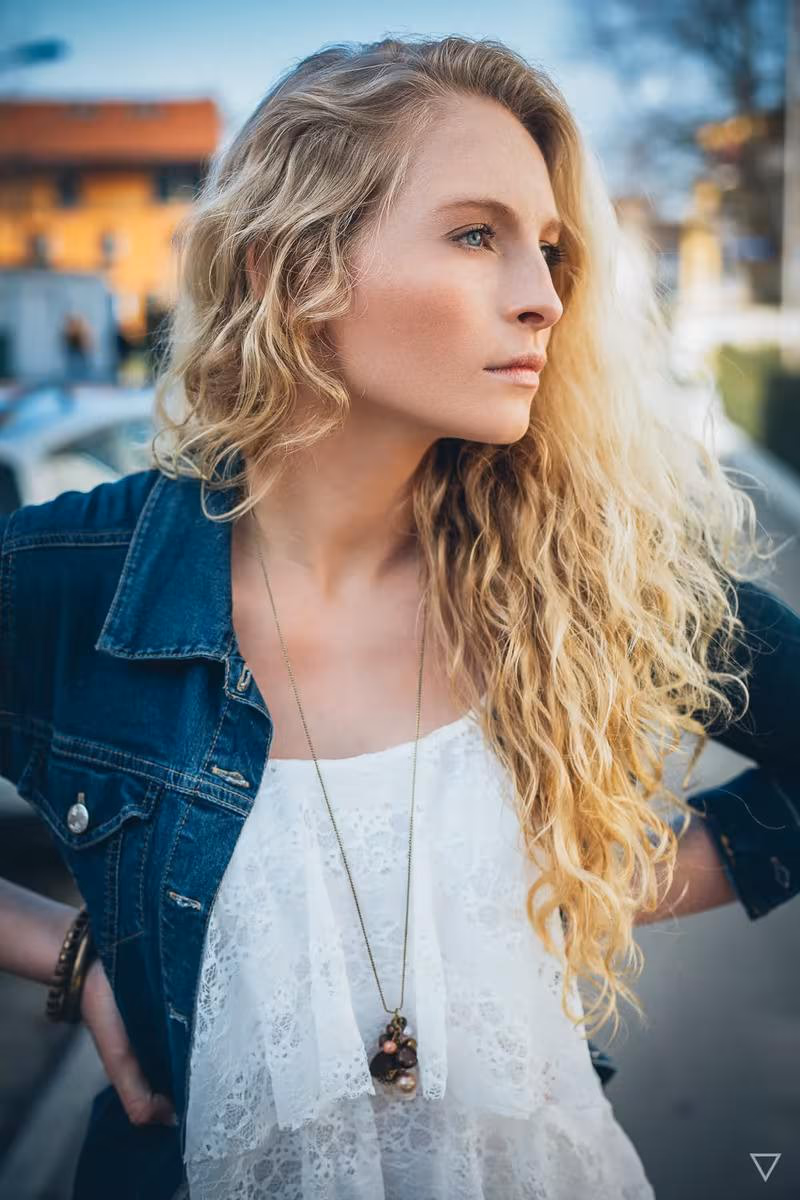 Lifestyle: Blonde Frau mit lockigem Haar in weißem Spitzenkleid und Jeansjacke im urbanen Raum.