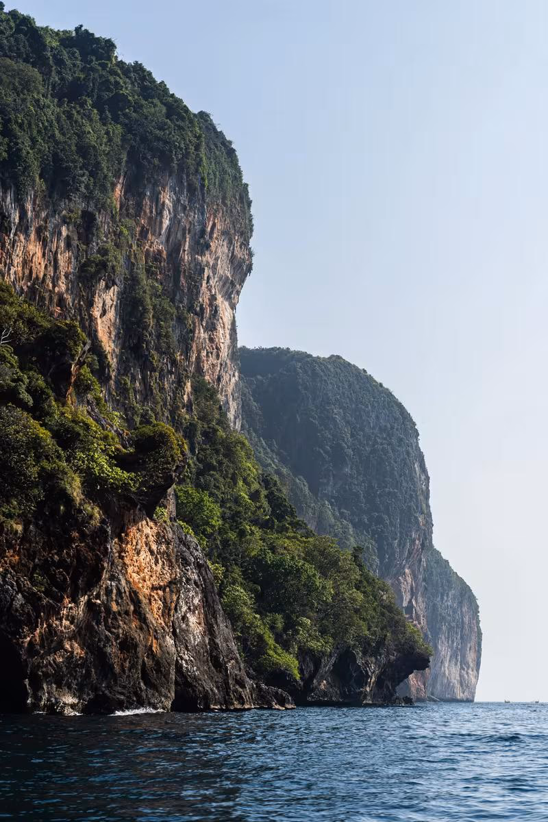 Thailand: Steile, bewaldete Klippen am Meer unter hellem Himmel.