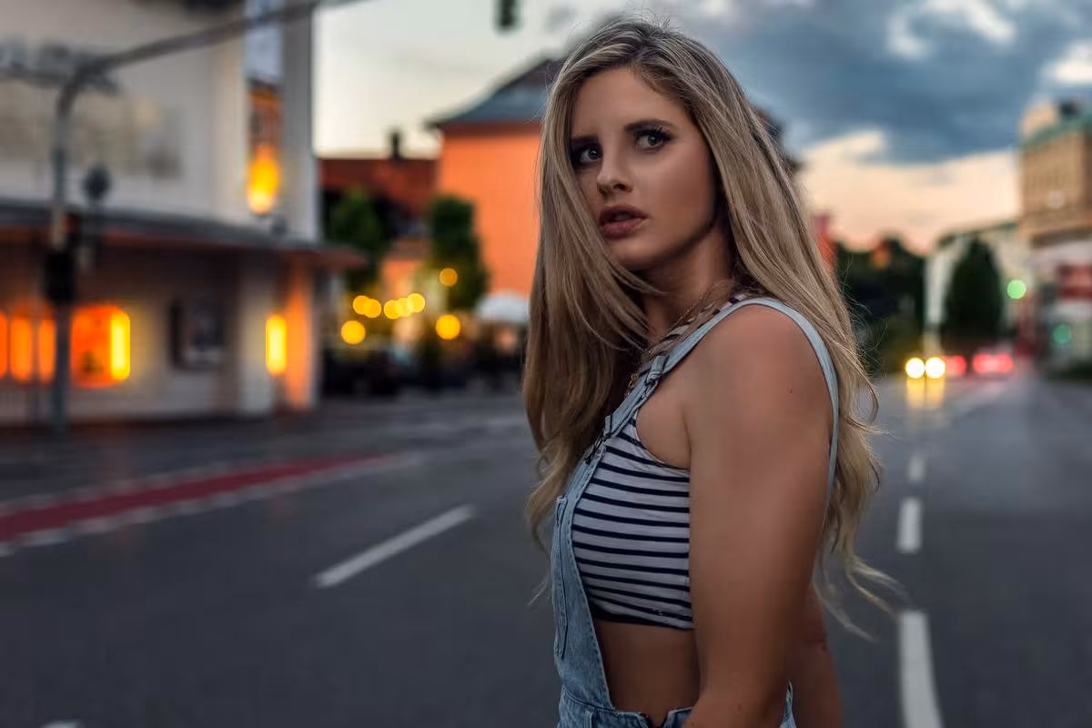 Lifestyle: Blonde Frau in gestreiftem Top und Latzhose auf einer Stadtstraße bei Dämmerung.