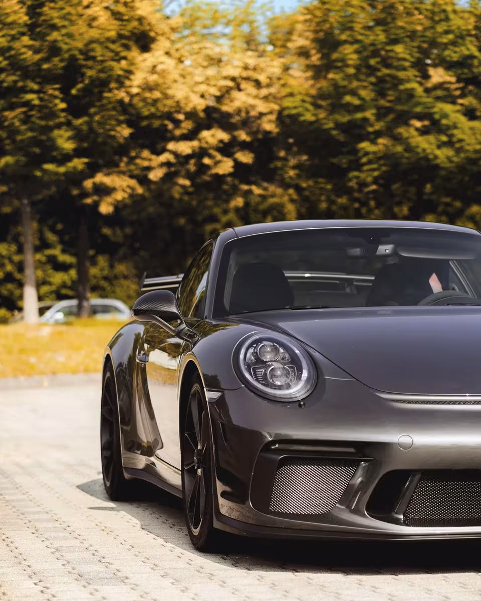 Audi: Schwarzer Porsche 911 GT3 mit Carbon-Details auf gepflasterter Fläche vor Bäumen.
