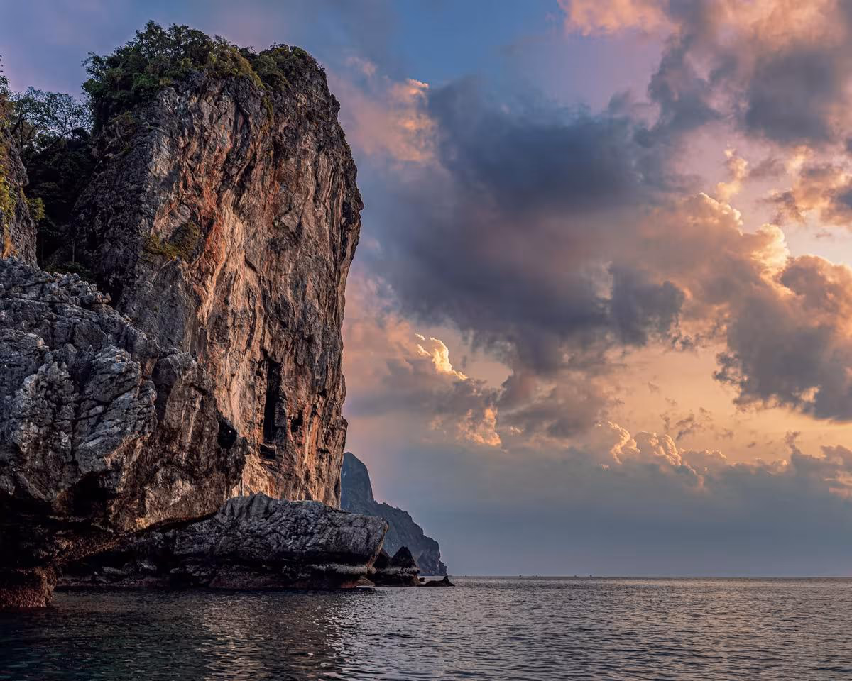 Thailand: Steile Felsklippe am Meer bei dramatischem Sonnenuntergang mit rosafarbenen Wolken.