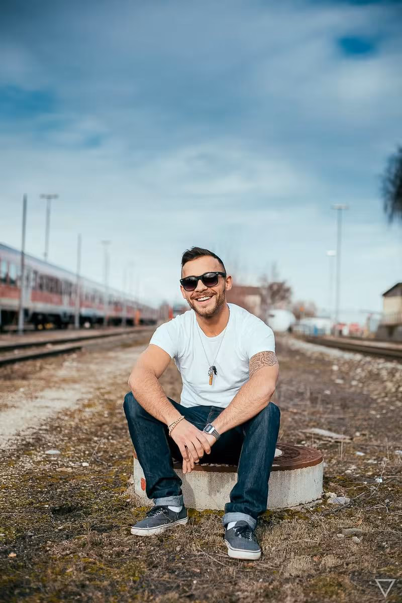 Lifestyle: Lachender Mann mit Sonnenbrille sitzt auf Betonblock an Gleisen.