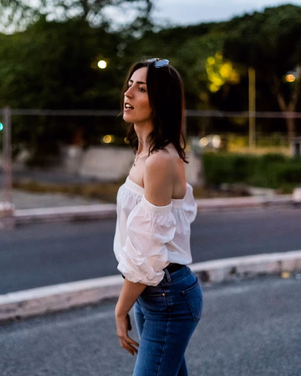 Lifestyle: Frau in weißem Off-Shoulder-Top und Jeans auf einer Straße bei Dämmerung.