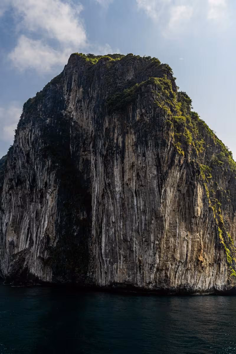 Thailand: Steile, bewachsene Felswand, die senkrecht aus dunklem Meerwasser aufragt.