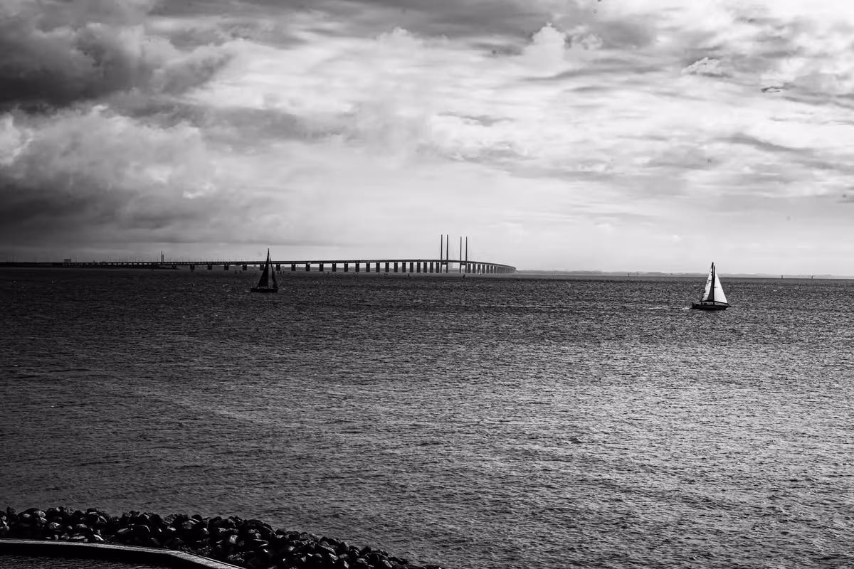 Copenhagen: Schwarz-Weiß-Aufnahme: Zwei Segelboote vor einer langen Brücke unter bewölktem Himmel.