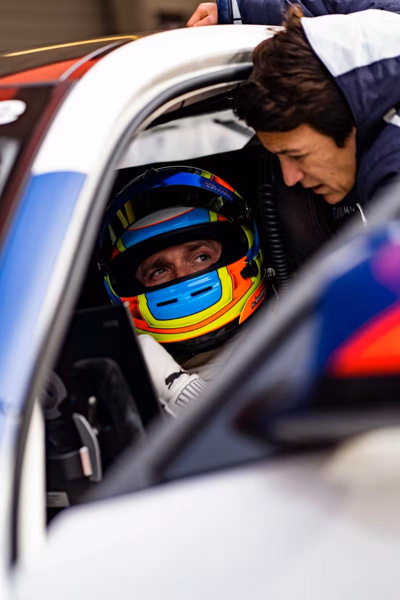 BMW Motorsport: Fahrer im bunten Helm im BMW-Renncockpit, Mechaniker beugt sich hinein.