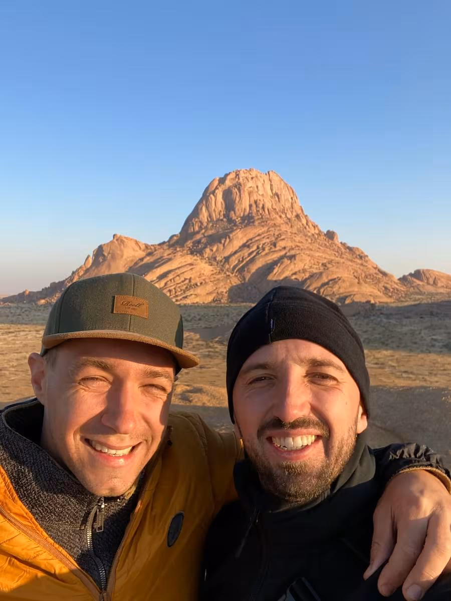 Fujifilm: Zwei Männer lächeln im Selfie vor einer felsigen Wüstenlandschaft bei Sonnenuntergang.