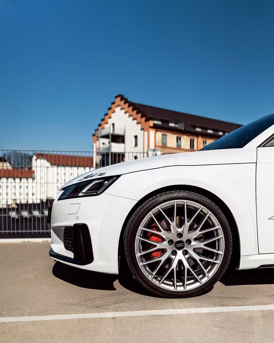 Vorderansicht eines weißen Audi mit silbernen Felgen und roten Bremssätteln vor einem Gebäude.