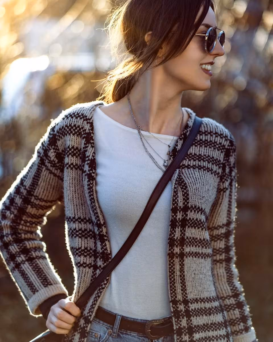 Lifestyle: Lachende Frau mit Sonnenbrille, gestricktem Cardigan und Lederriemen im warmen Licht.