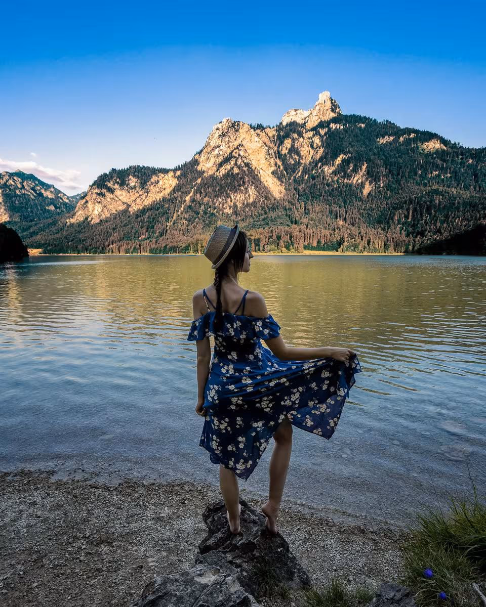 Lifestyle: Frau im blauen Kleid und Hut steht auf Felsen am Bergsee.