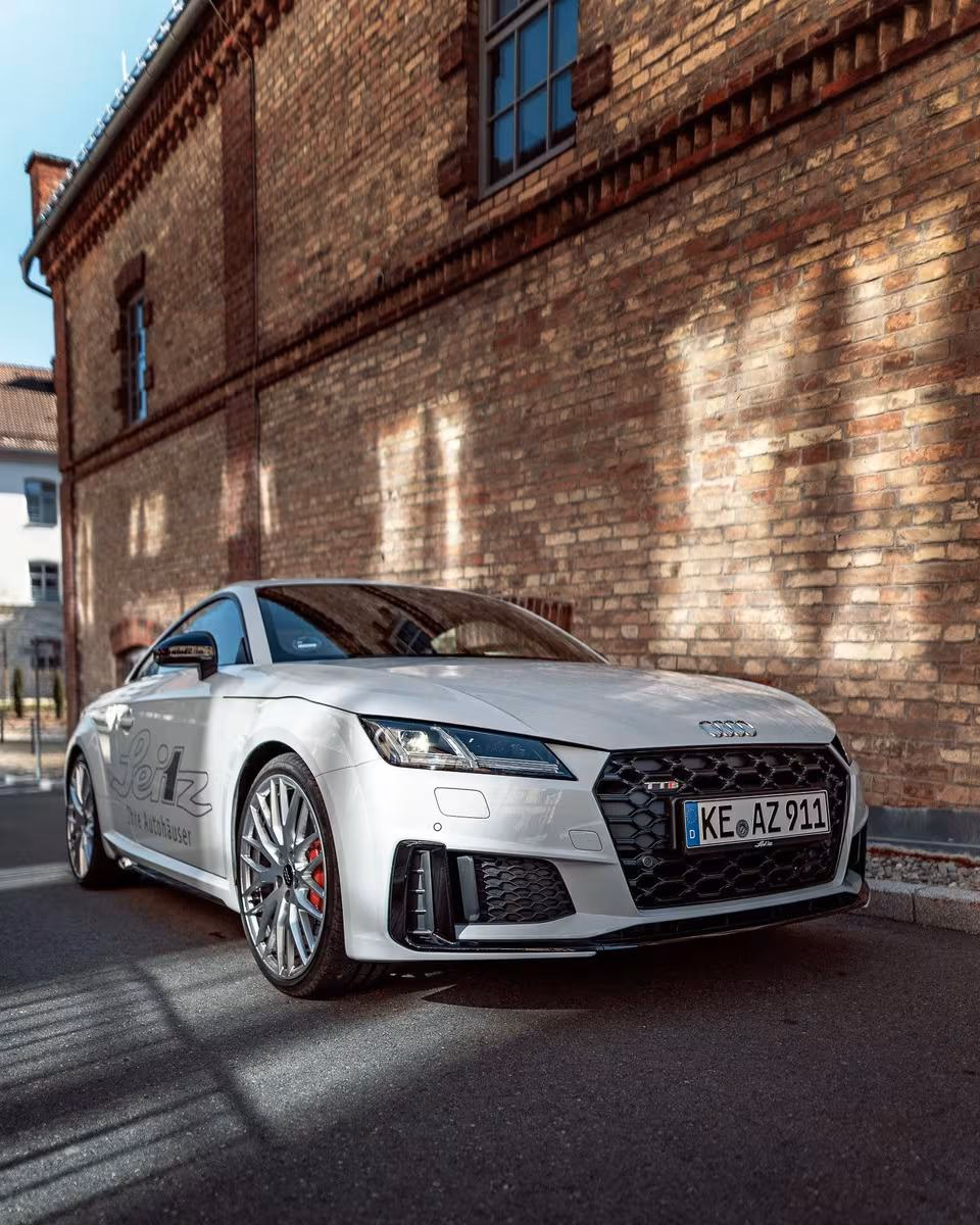 Silberner Audi TT S vor roter Ziegelwand mit Seitenschrift „Seiz Ihre Autohäuser“.