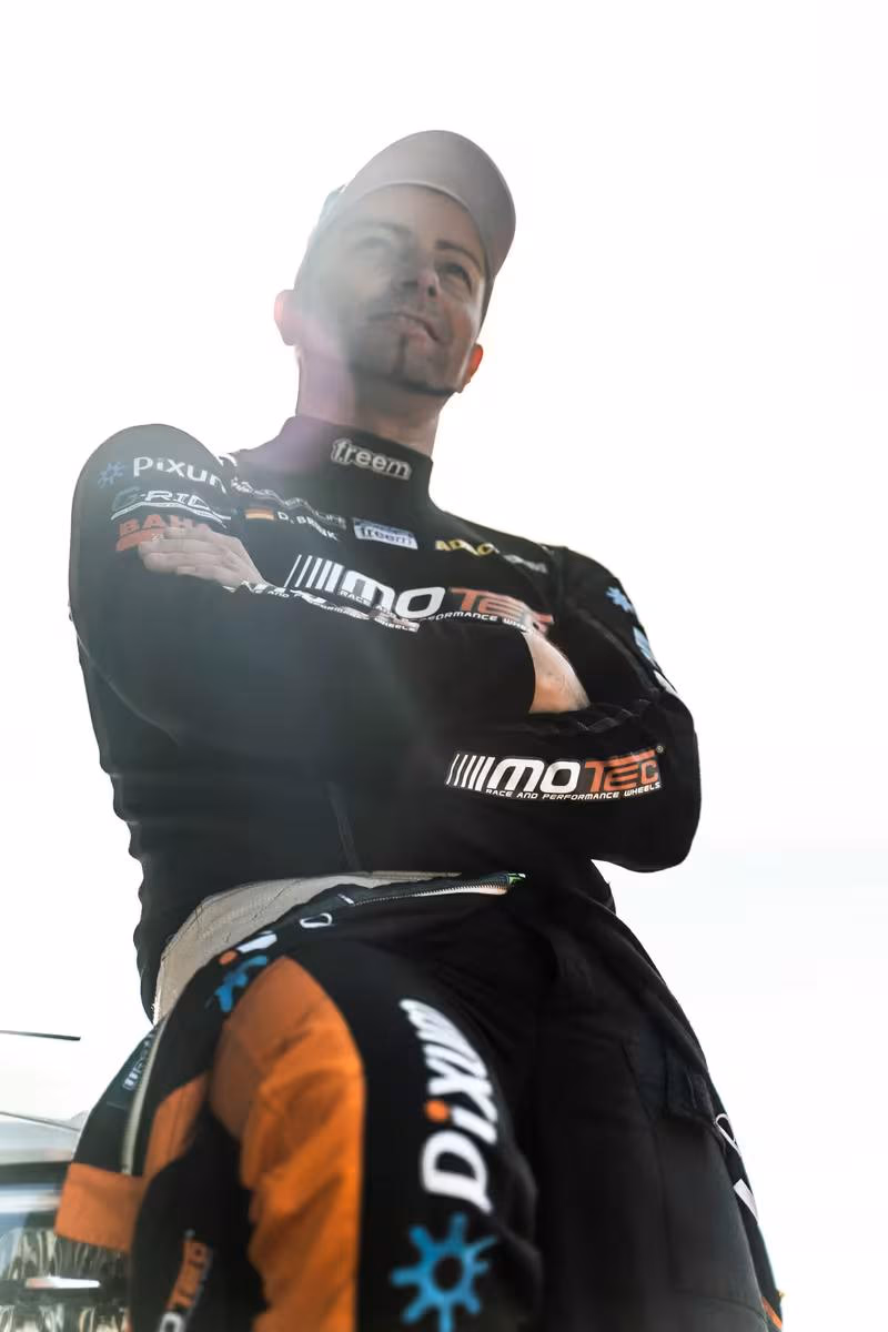 BMW Motorsport: Rennfahrer in schwarzem Overalls mit MoteC- und Pixum-Logos, Arme verschränkt.