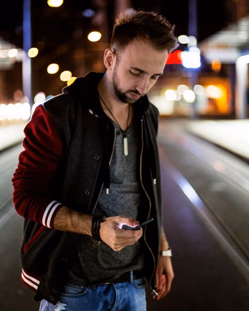 Lifestyle: Mann in Lederjacke blickt nachts bei Stadtlichtern auf sein Handy.