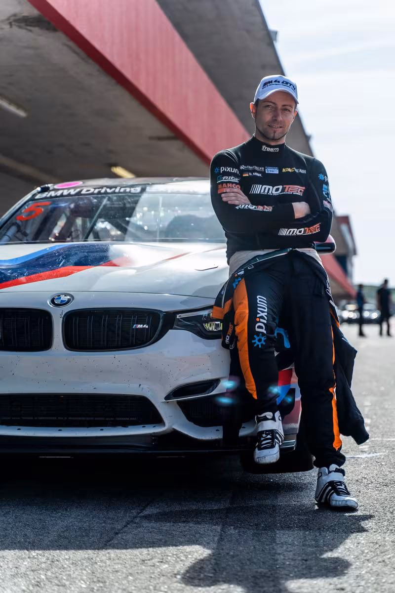 BMW Motorsport: Rennfahrer in schwarzem Overalls lehnt lässig an einem weißen BMW M4 GT3 Rennwagen.