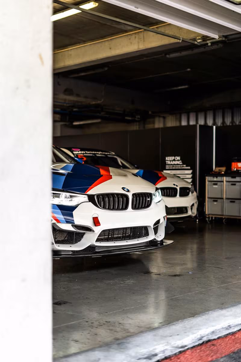 Zwei BMW M6 GT3 Rennautos mit Motorsport-Lackierung im Innenraum eines Garagen-Boxenbereichs.
